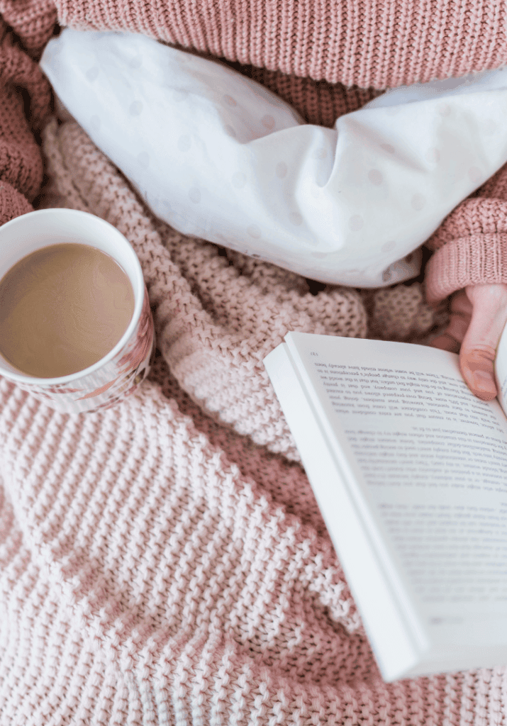 a woman holding her coffee in one hand and a book in another while wrapped in a blanket. 