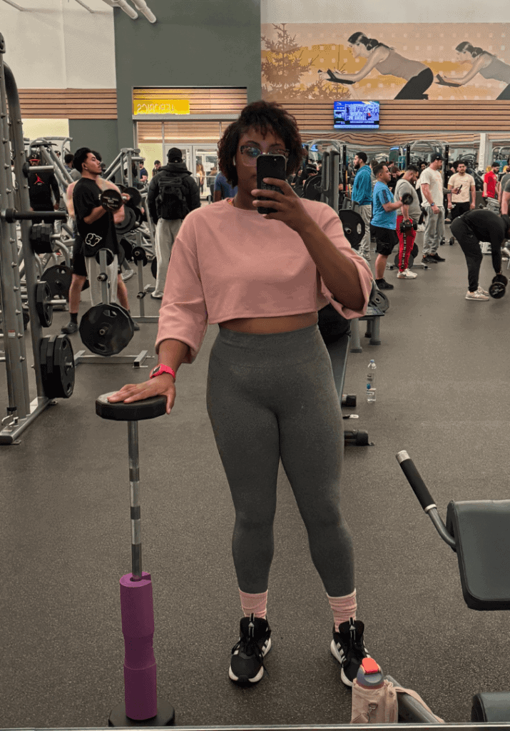 Brianna Imani in a pink top and grey leggings showing her fitness transformation in a gym mirror picture. 
