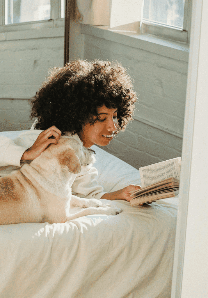 black woman reading a book in the summer on her bed with her dog.