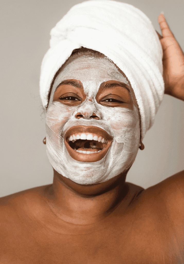 a smiling black woman with a face mask and a spa towel wrapped around her head. 